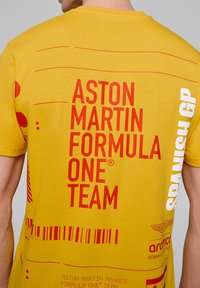 Żółta bawełniana koszulka z wyraźnym czerwonym i białym nadrukiem, zawierająca tekst "Aston Martin Formula One Team" oraz akcenty wzorów.