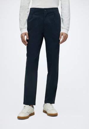 Homme portant un pantalon droit bleu marine foncé, un haut à manches longues blanc et des baskets blanches à semelles marron, debout sur un fond uni.