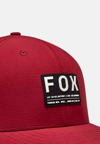 Gorra roja de tela texturizada con un parche rectangular negro que lleva las letras "FOX" en blanco. Líneas limpias y forma redondeada.