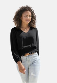Schwarze V-Ausschnitt-Bluse mit langen Ärmeln und gerafftem Saum, kombiniert mit distressten hellblauen Jeans und einem hellbraunen Gürtel.