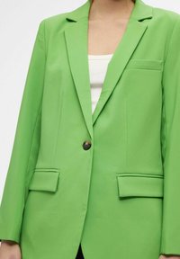 Heldergroene blazer met een getailleerde pasvorm, enkel knoopsluiting, uitgesneden revers en twee voorzakken. Gladde, gestructureerde stofafwerking.