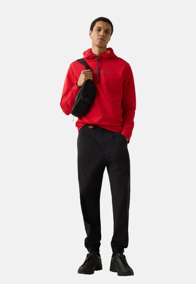 Jeune homme portant un sweat à capuche rouge, un pantalon de jogging noir, des baskets épaisses noires et un sac bandoulière noir, debout devant un fond blanc uni.