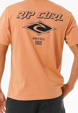 Rückansicht einer Person, die ein pfirsichfarbenes T-Shirt mit schwarzem Rip Curl-Logo, einer stilisierten Wellen-Grafik und dem Text "Born at Bells 1969." trägt.