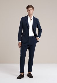 Mornarskoplavi muški odijelo s prilagođenim dizajnom, zatvaranje na jedan gumb, odgovarajuće hlače i bijela košulja. Tamne kožne cipele na slip-on.