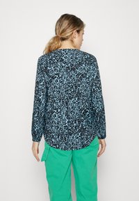 Langermet bluse i et lyseblått stoff med et svart blomstermønster. Har en rund kant og en myk tekstur. Kombinert med grønne bukser.