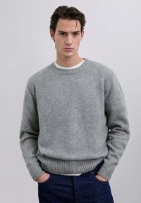 Suéter gris de punto con cuello redondo, puños y dobladillo acanalados. Usado sobre una camisa blanca, tiene un acabado suave y texturizado.