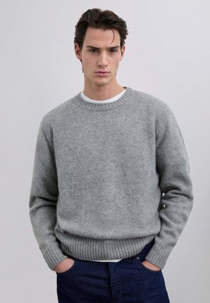 Suéter gris de punto con cuello redondo, puños y dobladillo acanalados. Usado sobre una camisa blanca, tiene un acabado suave y texturizado.