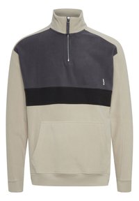 Sudadera de forro polar en beige y gris oscuro con cuello de media cremallera, bolsillo frontal y banda de acento negra en el pecho. Textura suave.