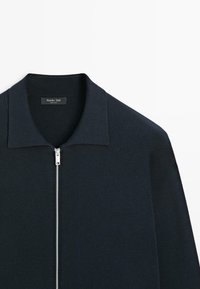 Navy zip-up sweater with a flat collar, ribbed texture, and silver zipper. Features raglan sleeves and a discreet brand label inside the collar.