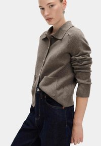 Brązowy kardigan z wełny z kołnierzykiem, zapięciem na guziki i przednimi kieszeniami. Rękawy wykończone ściągaczem i luźny krój. Nosi ciemne dżinsy.