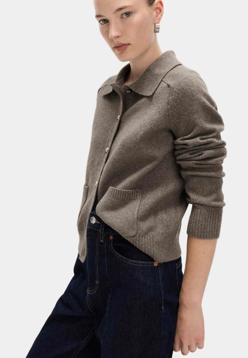 Brązowy kardigan z wełny z kołnierzykiem, zapięciem na guziki i przednimi kieszeniami. Rękawy wykończone ściągaczem i luźny krój. Nosi ciemne dżinsy.