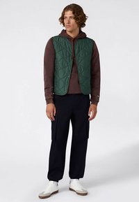 Groene doorgestikte bodywarmer over een bruine hoodie, gecombineerd met marineblauwe cargobroeken en witte sneakers. Voorzien van drukknoopsluitingen en een opstaande kraag.