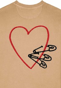 Sweatshirt beige avec un grand cœur rouge brodé et trois épingles de sûreté noires, avec des détails de couture texturés sur le tissu.