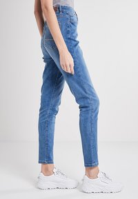 Zilā denim bikses ar šauru piegriezumu, augstu jostasvietu, nelielu nodilumu un izšūtām akcijām. Apvienotas ar masīvām baltām kedām.