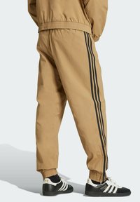 Tan track pants with black side stripes, elastic waistband and cuffs, featuring zippered ankles. Paired with black and white sneakers.