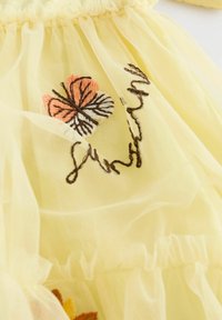 Tissu en tulle jaune avec des volants empilés. Comporte un papillon brodé en orange et blanc ainsi qu'un détail textuel en fil marron foncé.