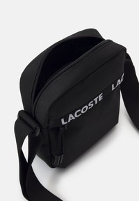 Borsa a tracolla nera in nylon con chiusura a zip, tracolla regolabile e logo bianco "LACOSTE" sul pannello anteriore. Texture liscia.