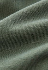 Gros plan sur un tissu tricoté doux et vert montrant des coutures fines et régulières ainsi que des plis délicats créant texture et profondeur.