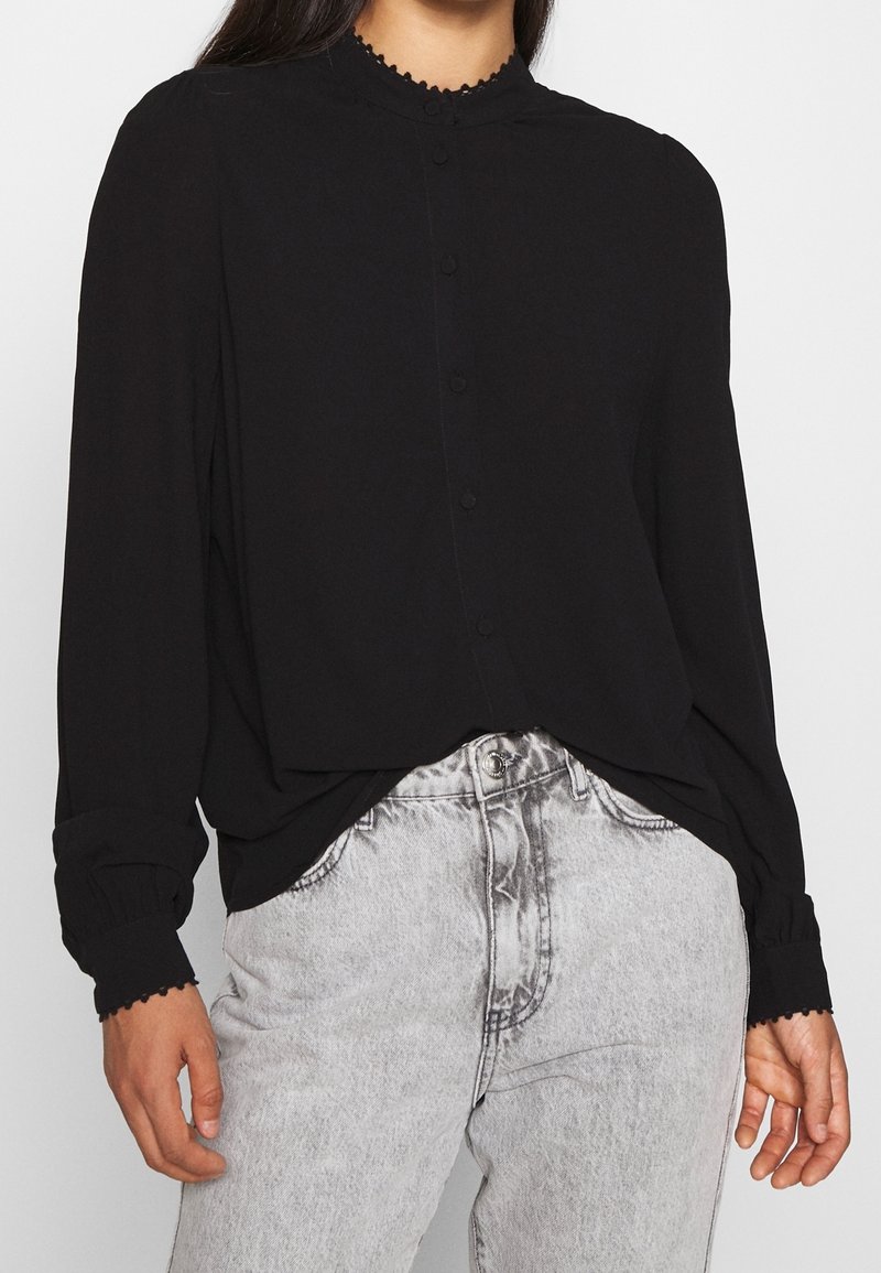 Femme portant un chemisier noir à manches longues boutonné avec des poignets et un col ornés de dentelle, associé à un jean taille haute gris clair.