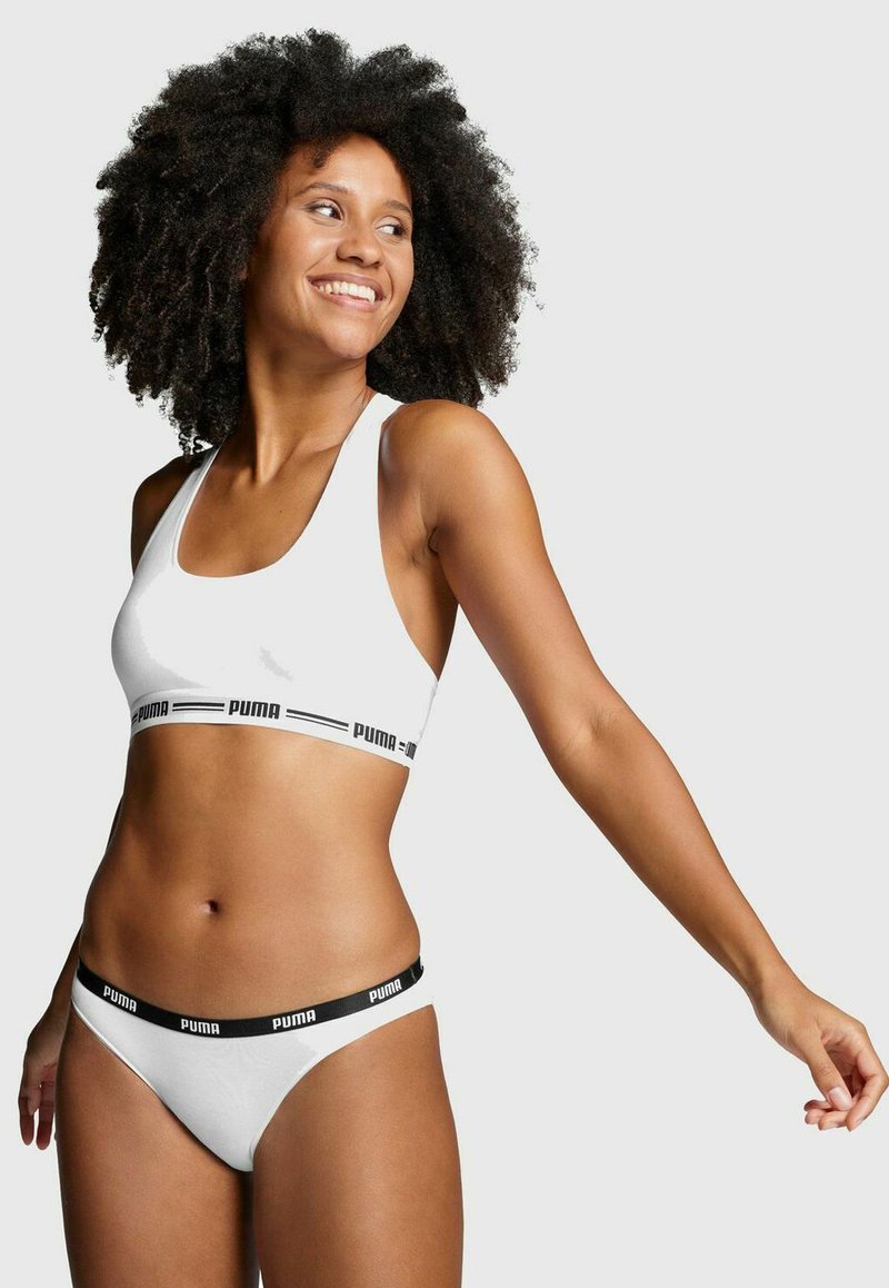 Weißer Sport-BH mit schwarzem Puma-Logo-Band, kombiniert mit weißen Bikini-Höschen, die einen kontrastierenden schwarzen Bund haben.