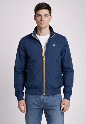 Młody mężczyzna zwrócony przodem, ubrany w granatową kurtkę na zamek błyskawiczny na białą koszulę i niebieskie dżinsy, stojący na jasnym, jednolitym tle.