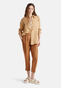 Světle hnědá lněná košile, bílý top a měděně zbarvené kalhoty se zúženým střihem a uvolněným střihem. Doplňuje béžové chunky sandály. Jednoduchý design.