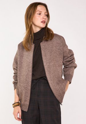 Giacca bomber in maglia marrone con polsini e orlo a costine, caratterizzata da una chiusura con zip. Indossata sopra un dolcevita scuro e abbinata a pantaloni a quadri.