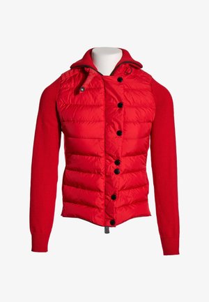 Rød jakke med vatteret front, sorte knapper, strikkede lange ærmer og en stående krave med lynlås og snøre.