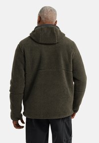 Fleecejacke in dunkelgrün mit einer weichen, strukturierten Oberfläche und einer enganliegenden Kapuze. Verfügt über eine Naht auf der Rückseite und seitliche Taschen.