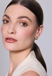 Złote kolczyki w kształcie obręczy z przekręconym wzorem, składające się z trzech nałożonych na siebie pasków, noszone przez osobę o jasnej karnacji i prostych włosach.