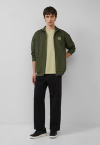 Olivgrüne Zip-Jacke mit seitlichen Taschen, kombiniert mit einem hellgelben T-Shirt und schwarzen weiten Hosen. Schwarze Sneaker runden das Outfit ab.