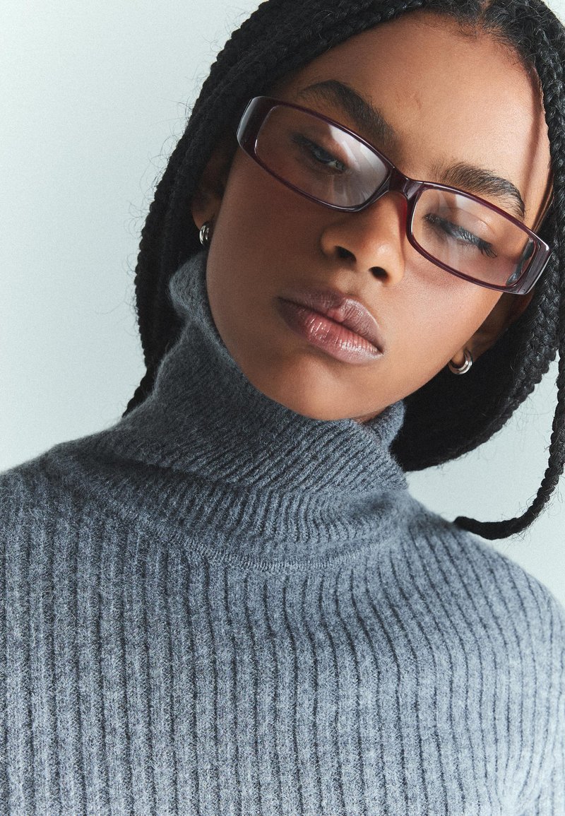 Siv škrbasta turtleneck pulover iz mehkega materiala, ki se ponaša z visokim ovratnikom in teksturiranim pletenjem, nosi se z temnimi očali.
