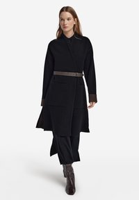 Schwarzer, langer Wollmantel mit Wickeldesign, zwei Vordertaschen und einem braunen Gürtel. Die Manschetten haben eine kontrastierende braune Textur.