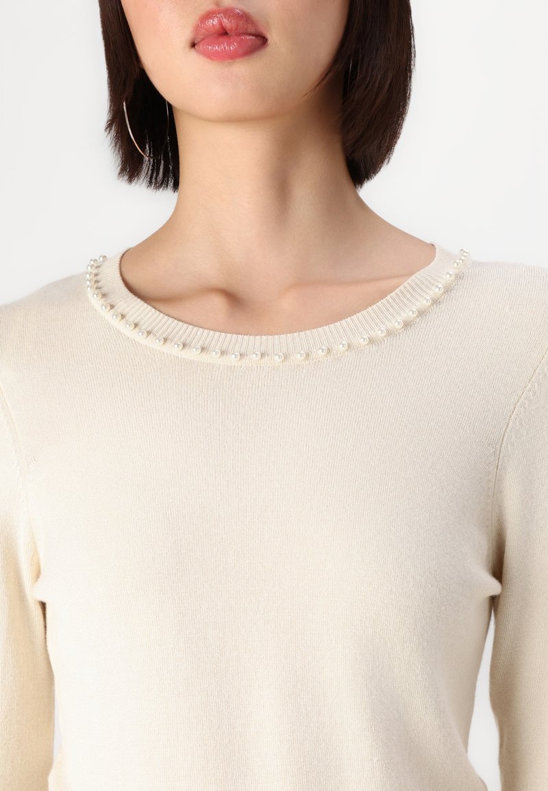 Maglione beige chiaro con una texture morbida, caratterizzato da un collo rotondo adornato da una fila di piccole perle bianche lungo il colletto.