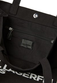 Borsa tote in tela nera con una tasca interna con zip, un'etichetta in pelle nera e una chiusura a scatto nella parte superiore. Grande testo bianco all'esterno.