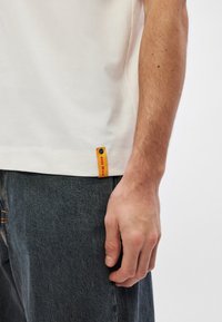 Λευκό βαμβακερό t-shirt με στρογγυλή λαιμόκοψη, κοντά μανίκια και μια μικρή πορτοκαλί ετικέτα με το "BOSS" στο τελείωμα. �Ορατό μπλε τζιν παντελόνι.