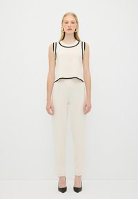 Top senza maniche beige chiaro con bordi neri, abbinato a pantaloni aderenti beige chiaro. Il modello indossa décolleté nere a punta. Design minimalista.
