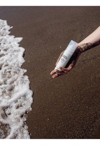 Main recouverte de sable mouillé tenant un flacon de crème solaire près des vagues mousseuses de l'océan sur une plage de sable.