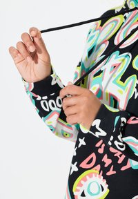 Sweatshirt à capuche avec un fond noir et des motifs graphiques colorés en rose, turquoise et blanc. Un cordon est tenu par une main.