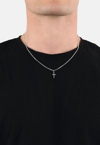 Mann trägt eine silberne Kettenkette mit einem kleinen Kreuzanhänger über einem schwarzen Rundhals-Shirt vor einem weißen Hintergrund.