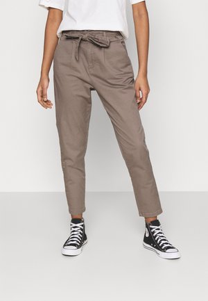 Kobieta w beżowych spodniach z wysokim stanem typu paperbag związana paskiem i czarnych wysokich sneakersach, stojąca na jasnym, jednolitym tle.