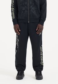 Svart huvtröja med intrikat svart grafiskt mönster; matchande svarta sweatpants med guldaccenter och en stiliserad drakeprint på benet.