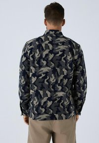 Man die zich omdraait en een langefysieke camouflagedruk met grijs en marineblauw draagt, gecombineerd met lichtbruine broek.