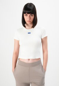 Jonge vrouw met steil donker haar, gekleed in een witte Hugo t-shirt met korte mouwen en beige broek, met haar handen in haar zakken, tegen een effen achtergrond.