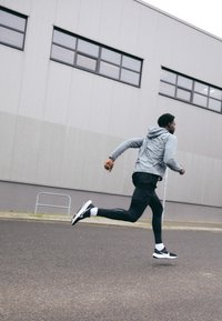 Idrettsklær inkluderer en grå hettegenser, svarte shorts og svarte tights. Løperen har på seg svarte og hvite joggesko, fanget midt i steget.