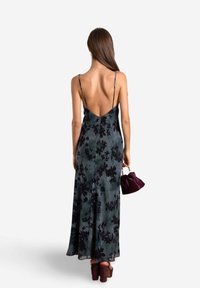 Langes, rückenfreies Kleid in Dunkelgrün mit schwarzem Blumenmuster, dünnen Trägern und einer fließenden Silhouette; kombiniert mit einer Handtasche in Pflaumenfarbe und hohen Schuhen.
