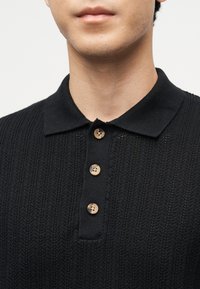 Homme portant un polo noir en maille avec trois boutons marron, montré du cou à la partie supérieure de la poitrine.