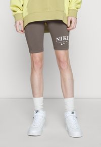 Bruna sportshorts med "NIKE"-logotyp i vitt, kombinerat med vita sneakers och krämfärgade strumpor. Bakgrunden är neutral grå.