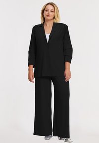 Zwarte blazer en wijde broek gemaakt van lichtgewicht stof. De blazer heeft een gelapde kraag en twee knopen. Zilveren schoenen accentueren de outfit.