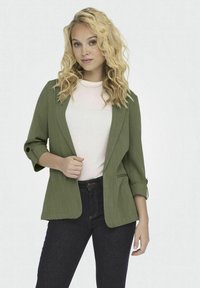 Blazer verde oliva con textura, de corte abierto y mangas tres cuartos. Combinado con una camisa blanca y jeans oscuros.